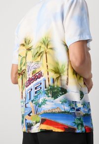 Farverig kortærmet skjorte med et strandmotiv, der inkluderer palmer, et vintage hotelskilt og levende nuancer af blå og gul. Glat stofstruktur.