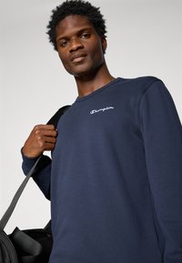 Navy langærmet skjorte lavet af bomuld, med hvidt broderet "Champion" logo, rund halsudskæring og manchetter. Sort taske holdes af modellen.