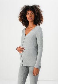 Cardigan de maternitate gri deschis, cu textură ripată, guler în V, închidere cu nasturi și mâneci lungi, asortat cu blugi gri mulați. Textură moale și ajustare confortabilă.