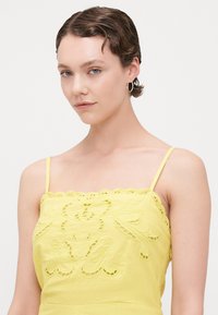 Camiseta amarilla con diseño floral bordado y borde festoneado, con tirantes finos y una tela texturizada.