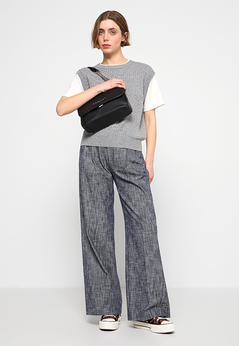 Donna in gilet di maglia grigia, camicia bianca a maniche corte, pantaloni blu a gamba larga, sneakers con stampa leopardata, che tiene una borsa nera a tracolla sul petto.