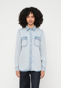 Light blue denim shirt with long sleeves, button-up front, two chest pockets, and contrasting blue stitching. Smooth texture, relaxed fit.