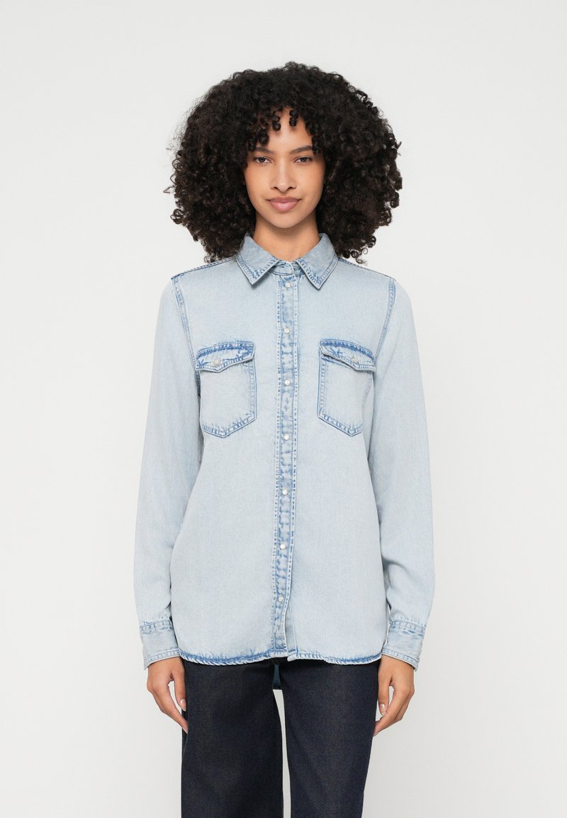 Light blue denim shirt with long sleeves, button-up front, two chest pockets, and contrasting blue stitching. Smooth texture, relaxed fit.
