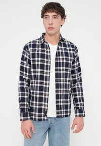 Πουκάμισο από φανέλα με καρό μοτίβο σε ναυτικό μπλε και άσπρο, με κόκκινες λεπτομέρειες, μακριά μανίκια, γιακά κουμπωτό και άνετη εφαρμογή. Φορεμένο πάνω από ένα άσπρο t-shirt.