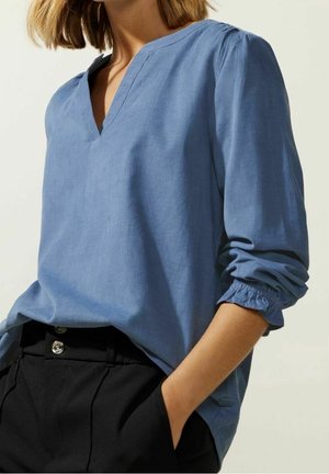 Vrouw met een blauwe blouse met lange mouwen en V-hals met geplooide manchetten en een zwarte broek met hoge taille, hand in de zak, tegen een lichte achtergrond.