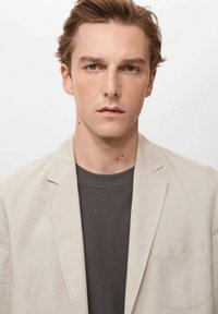 Blazer de lino color beige claro sobre una camiseta gris oscura, con cuello de muesca y un solo bolsillo en el pecho. Textura suave, diseño minimalista.