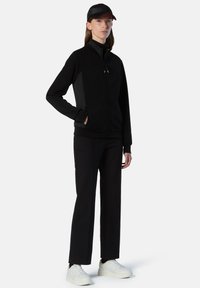 Chaqueta negra con cremallera y paneles laterales, combinada con pantalones negros y zapatillas blancas. La gorra coincide con el color de la chaqueta.