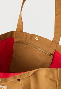 Καφέ τσάντα ώμου από καμβά με κόκκινη επένδυση στο εσωτερικό, περιλαμβάνει εσωτερική τσέπη με φερμουάρ και κλείσιμο με κουμπί κοντά στην κορυφή της.