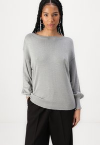 Suéter cinza claro, de corte solto, com um decote largo e mangas longas. Tecido texturizado com um brilho subtil, combinado com calças pretas.