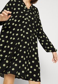 Musta mekko vihreillä pallokuoseilla, jossa on kaulus, pitkät hihat ja löysä leikkaus, joka levenee rinnasta. Napit kaula-aukossa.