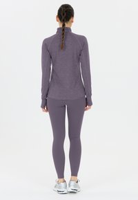 Kvinna med flätat hår som bär en matchande lila träningsöverdel och leggings, står vänd bort mot en vit bakgrund.