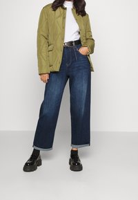 Quiltad olivgrön jacka med tryckknappar, vit polotröja, högmidjade mörkblå jeans med uppvikt nederkant, och svarta lackskor.