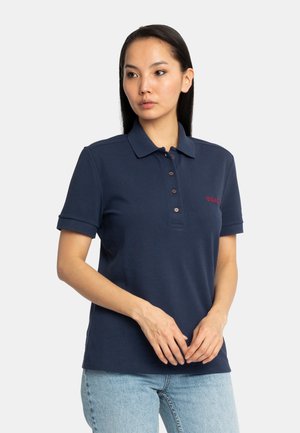 Femme portant un polo bleu marine à manches courtes et un jean bleu clair, debout devant un fond clair uni.