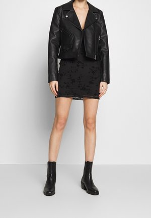 Schwarze Lederbikerjacke mit silberner Hardware, kombiniert mit einem figurbetonten schwarzen Kleid mit Blumenstickerei und knöchellangen schwarzen Stiefeln.