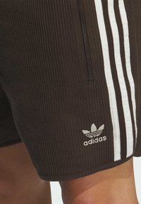 Hnědé žebrované šortky s bílými postranními pruhy, zipovou kapsou a vyšitým logem adidas na lemu. Tkanina má texturovaný povrch.