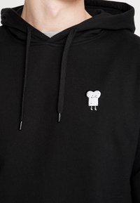 Felpa nera realizzata in tessuto morbido, con un piccolo personaggio di pane in stile fumetto ricamato sul lato sinistro del petto. Design semplice, modello a pullover.