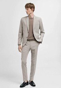 Beige skræddersyet jakkesæt med et subtilt mønster, parret med en brun crew-neck sweater og sorte lædersko. Slim fit, glat stoftekstur.