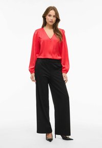 Blusa de satén roja con escote en V y mangas largas, combinada con pantalones anchos negros y tacones negros puntiagudos con acentos dorados.