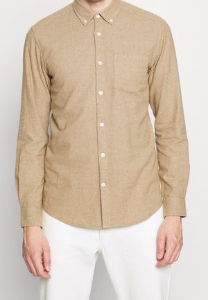 Hombre que lleva una camisa beige de manga larga abotonada con bolsillo en el pecho y pantalones blancos, de pie con las manos relajadas a los lados.