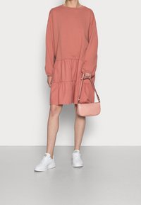 Vaaleanpunainen pitkähihainen college-mekko, jossa on kerroksellinen röyhelöhelma, yhdistettynä pieneen, kohotettuun vaaleanpunaiseen käsilaukkuun ja valkoisiin lenkkareihin.