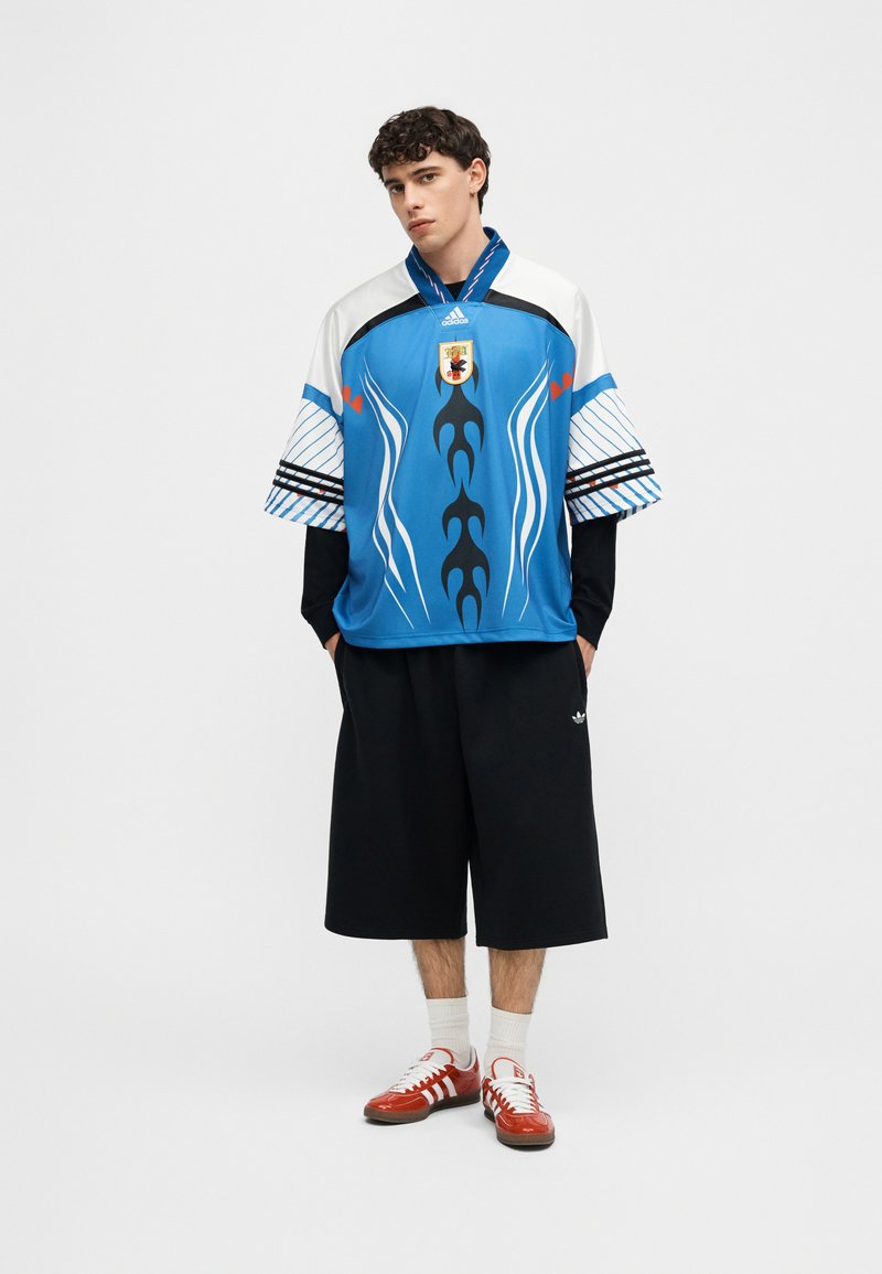 Jeune homme portant un maillot de sport bleu avec un motif graphique, un short large noir, des chaussettes blanches et des baskets rouges et blanches, debout devant un fond uni.