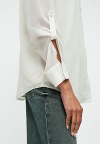 Kerge, läbipaistev valge pluus, millel on pikad varrukad, keeratud mansetid nupukestega ja sirge alläär, koos tuhmande sinise teksapükstega.