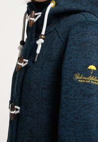 Blå texturerad hoodie med hög krage, vita dragsko, träknoppar och gult logotyp på sidan. Har stickad tyg och detaljer.