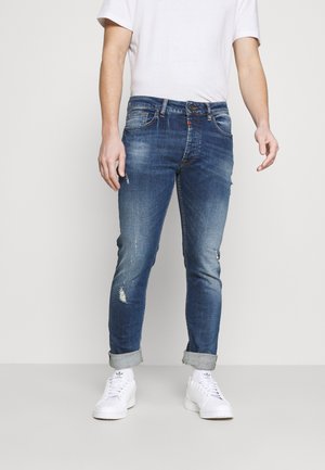 Schmal geschnittene blaue Denim-Jeans mit ausgefransten Details, fünf Taschen, Gürtelschlaufen und einem ausgewaschenen Finish. Kombiniert mit einem weißen T-Shirt und Sneakers.