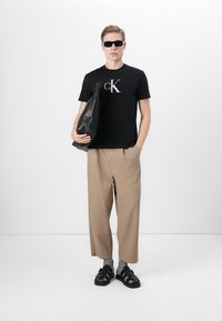 Zwarte katoenen T-shirt met zilver logo, losse beige broek en zwarte sandalen. Model houdt een grote zwarte leren tas vast. Donkere zonnebril gedragen.