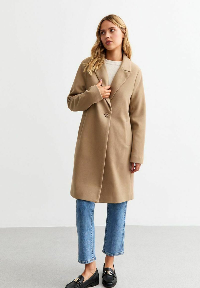 Beige lange jas met notch-kraag, enkele knoopsluiting, zijzakken en een zachte textuur, gecombineerd met lichtblauwe jeans en zwarte loafers.
