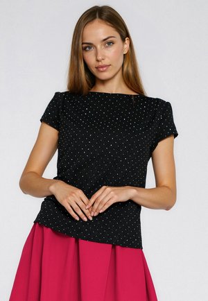 Kobieta o długich włosach, ubrana w czarną bluzkę z krótkim rękawem w drobne białe kropki oraz jaskrawo różową spódnicę, stojąca na jednolitym tle.