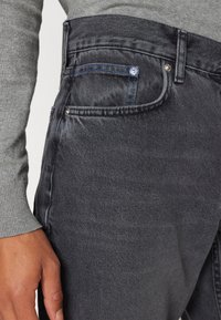 Jeans in denim grigio scuro con un taglio classico, caratterizzati da una leggera scoloritura, accenti di cuciture blu e tasche standard. La texture appare morbida e usurata.