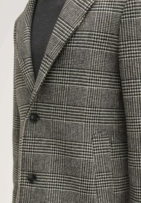 Hahnentrittmuster-Blazer in Schwarz und Weiß, mit einem Reverskragen, zwei schwarzen Knöpfen und einer einzelnen Brusttasche auf der linken Seite.