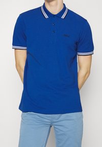 Blå pikétröja i bomull, med ribbad krage med vita ränder, treknapprad och en liten broderad logotyp på bröstet.