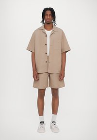 Hellbraunes Kurzarmhemd mit Knöpfen und Tasche, kombiniert mit passenden Shorts. Trägt weiße Socken und Sneakers. Minimalistisches Design.