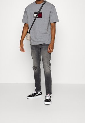 T-shirt en coton gris avec un logo imprimé, accents noir et rouge, associé à un jeans en denim gris déchiré et des baskets noir et blanc.