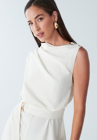 Witte, gestructureerde mouwloze top met een gedrapeerde halslijn. Heeft een getailleerde taille met een bijpassende riem en een opvallend zijwaarts knoopdetail.