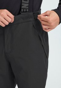 Black ski pants with a smooth, water-resistant fabric, featuring adjustable straps, a Velcro waistband, and side zip pockets.