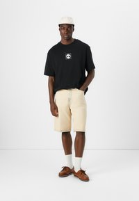 Svart oversized t-skjorte med logo, lysebeige denimshorts, kremfargede sokker og brune sko. Antrekket har en uformell, avslappet passform.