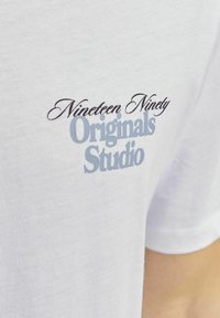 Valge puuvillane T-särk, mille esiküljel on sulgkirjas musta värvi "Nineteen Ninety" ja helesinine "Originals Studio".