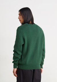 Πράσινη πλεκτή ζακέτα με ριμπ λαιμόκοψή και μανίκια. Διαθέτει μακριά μανίκια και χαλαρή εφαρμογή, υφή υφάσματος. Εμφανίζεται η πίσω όψη.