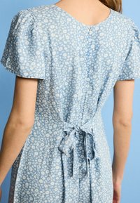 Mujer vestida con un vestido azul claro con estampado floral blanco, mangas cortas abullonadas, cremallera en la espalda y lazo de tela en la cintura.
