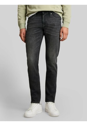 Mann trägt schwarze Slim-Fit-Jeans, weiße Sneakers und ein hellgrünes Sweatshirt und steht vor einem schlichten Hintergrund.