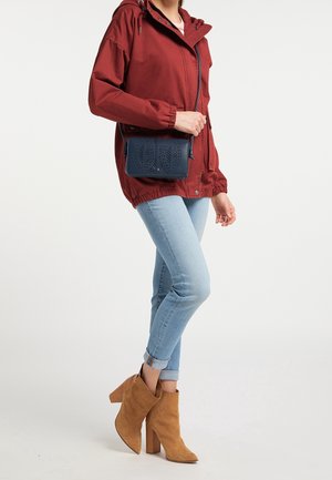 Woman wearing a rust-red jacket, light blue rolled-up jeans, tan suede high-heeled ankle boots, and holding a navy blue crossbody bag.