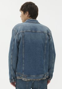 Vyras, vilkintis mėlyną džinsinę striukę ir džinsus, atsukęs nugarą, su languotu marškinėliu, matomu iš po striukės, prieš baltą vientisą foną.