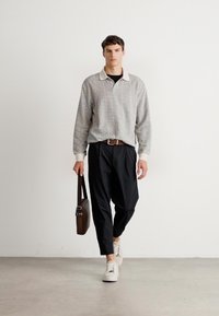 Man walking, wearing gray long-sleeve sweater, black pants, white sneakers, holding dark brown shoulder bag against plain white wall.