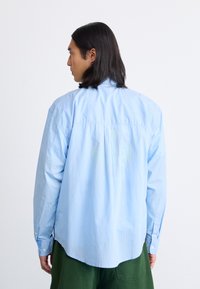 Camisa de manga larga a rayas azul claro, con puños abotonados y un dobladillo curvado. La tela parece suave con un ajuste relajado. Sin patrones o acentos prominentes.