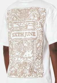 Camiseta blanca de algodón con un gráfico beige detallado en la parte de atrás que presenta diseños ornamentados y el texto "SIXTH JUNE" en un marco ovalado central.