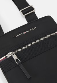 Borsa a tracolla nera in nylon con zip argentata, tracolla texturizzata e logo "TOMMY HILFIGER" in evidenza sul davanti.