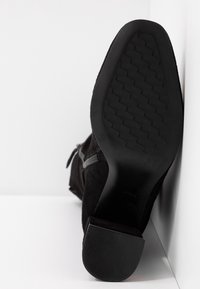 Botín negro de ante hasta el tobillo con una suela de goma texturizada, que presenta un zipper lateral y un tacón bajo y estratificado. Detalle de costura visible a lo largo de los bordes.